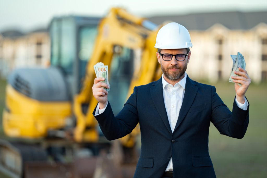 constuction worker in a suit wearing a hard had and holding money with a construction tractor excavator in the background