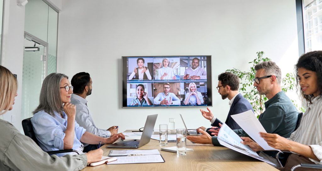 business meeting online in a converence room with a screen on the wall with virtual attendees